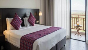 A hotel room with a large bed, a headboard, a nightstand, a bedside table, a window with curtains, and a balcony with a table and chair. - Donix Suites  (Njoro)