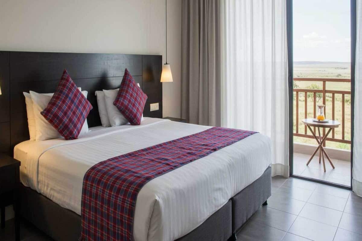 A hotel room with a large bed, a headboard, a nightstand, a bedside table, a window with curtains, and a balcony with a table and chair.