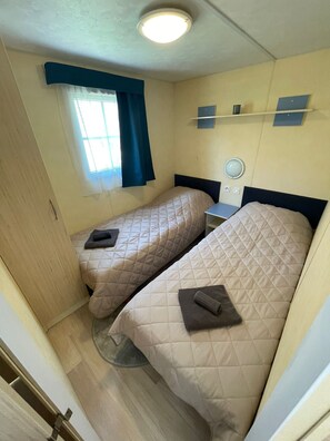 Ein kleiner, kompakter Raum mit zwei Einzelbetten, einem Fenster mit blauen Vorhängen, einem Nachttisch mit einer Lampe und einer Deckenleuchte.