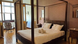 A four-poster bed with a wooden frame, a bedside table with a lamp, and a chair in a room with wooden flooring and a large window. - Vila Camélia - Suite Namorados (Porto)