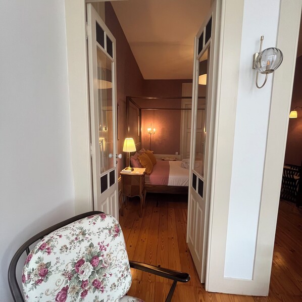 A narrow hallway leading to a bedroom with a bed, a bedside table with a lamp, and a chair with floral upholstery. - Vila Camélia - Suite Namorados (Porto)