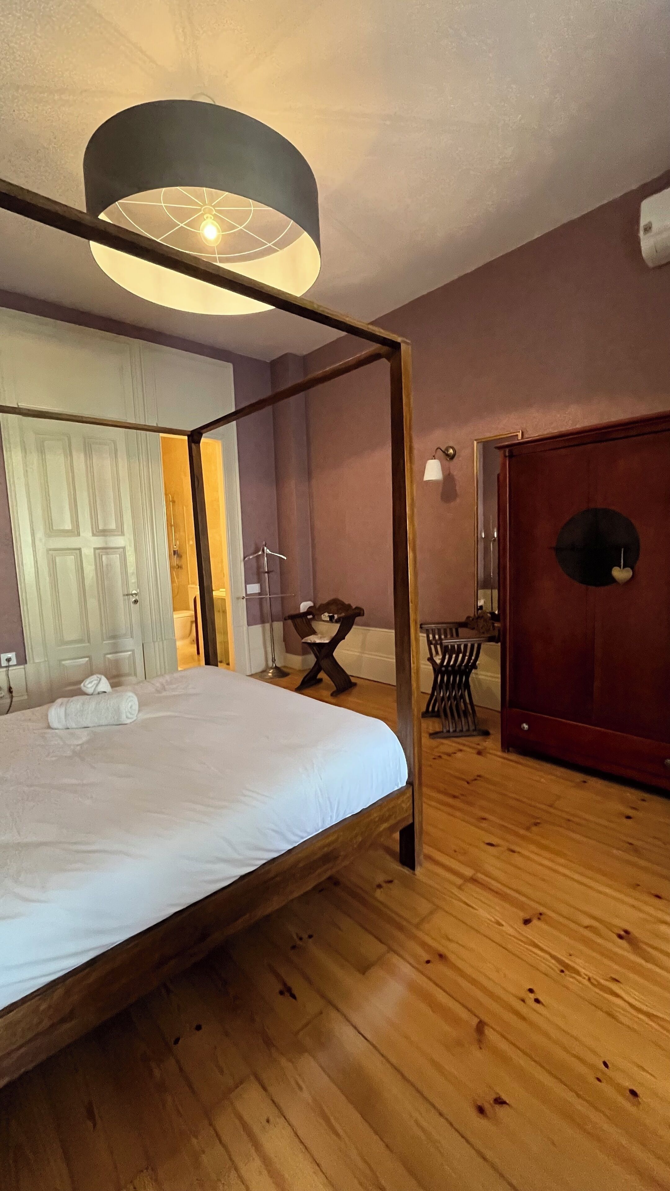 Een slaapkamer met een hemelbed, houten vloeren, een ronde spiegel en een garderobe.