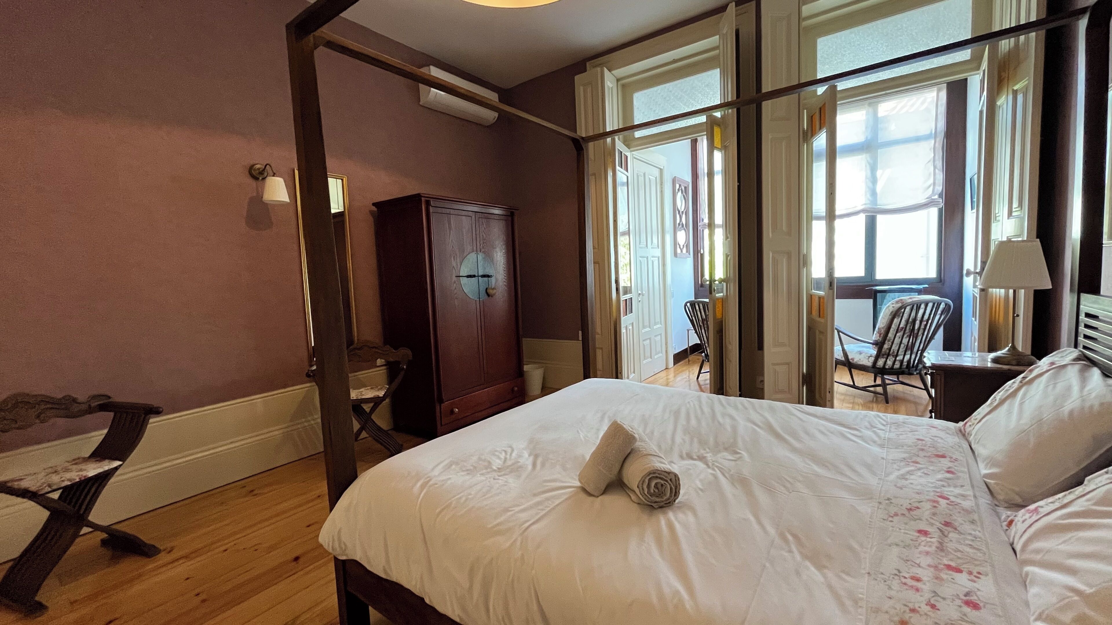 A bedroom with a bed, a wooden wardrobe, a chair, and a desk with a lamp.