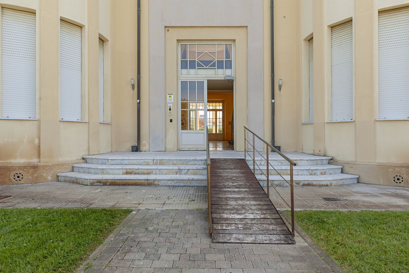 Ein Gebäudeeingang mit einer Treppe, die zu einer Tür führt, und flankiert von zwei großen Fenstern mit Rollläden.