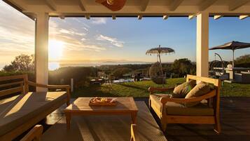 Une terrasse extérieure avec des meubles en bois, une table sur laquelle se trouve une pizza, et une vue sur le coucher de soleil se déclinant sur un paysage.