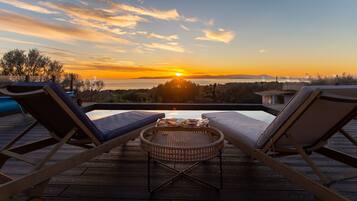 Une terrasse en bois avec deux chaises longues et une table en osier, offrant une vue imprenable sur le coucher du soleil.