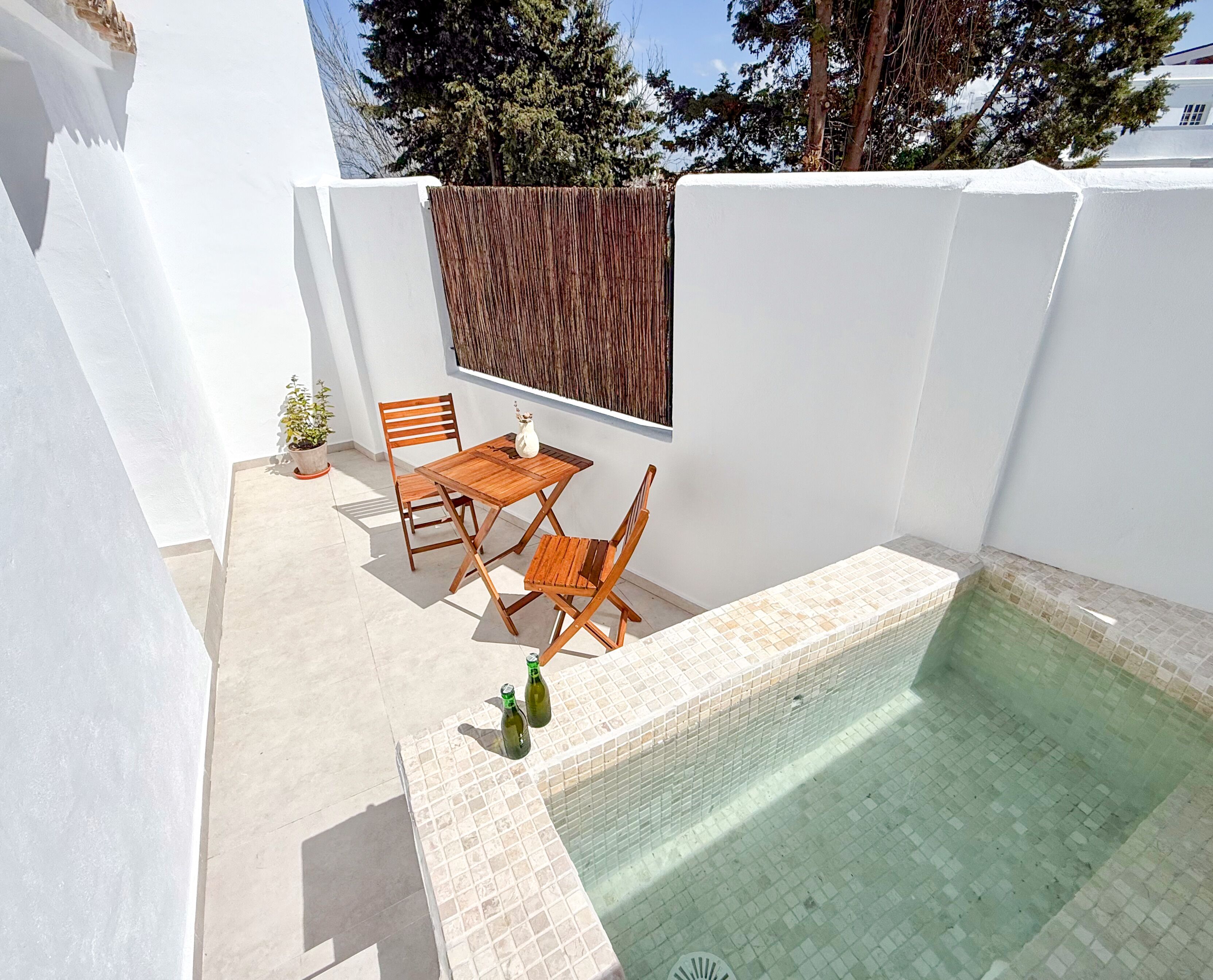 A pool area with a tiled edge, a wooden table and chairs, and a potted plant.