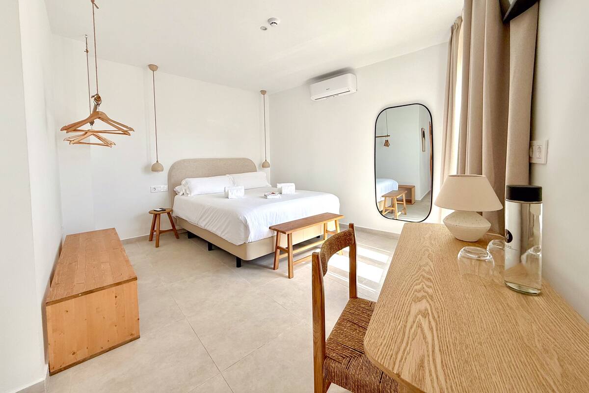 A bedroom with a bed, bedside table, chair, wooden bench, hanging light fixture, air conditioning unit, and a mirror.
