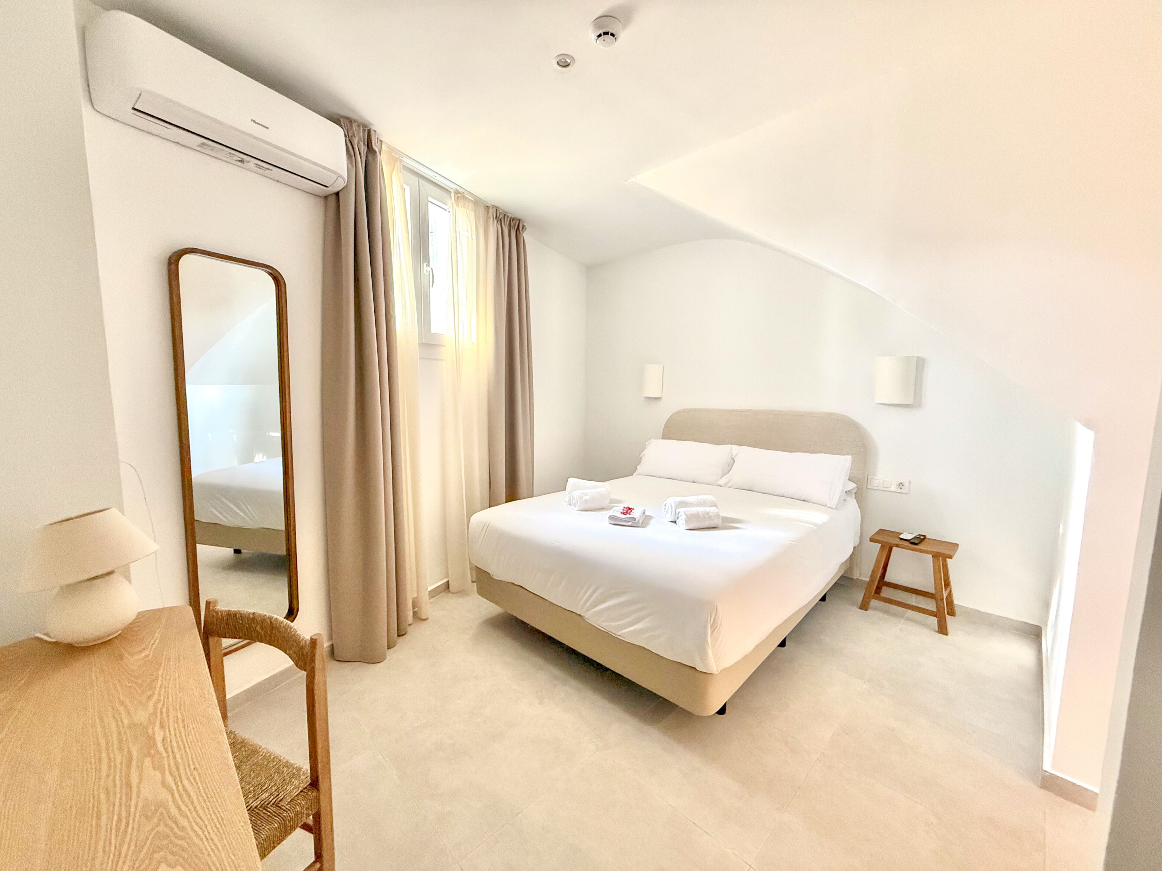 A hotel room with a bed, a desk, a chair, a mirror, and a small table.