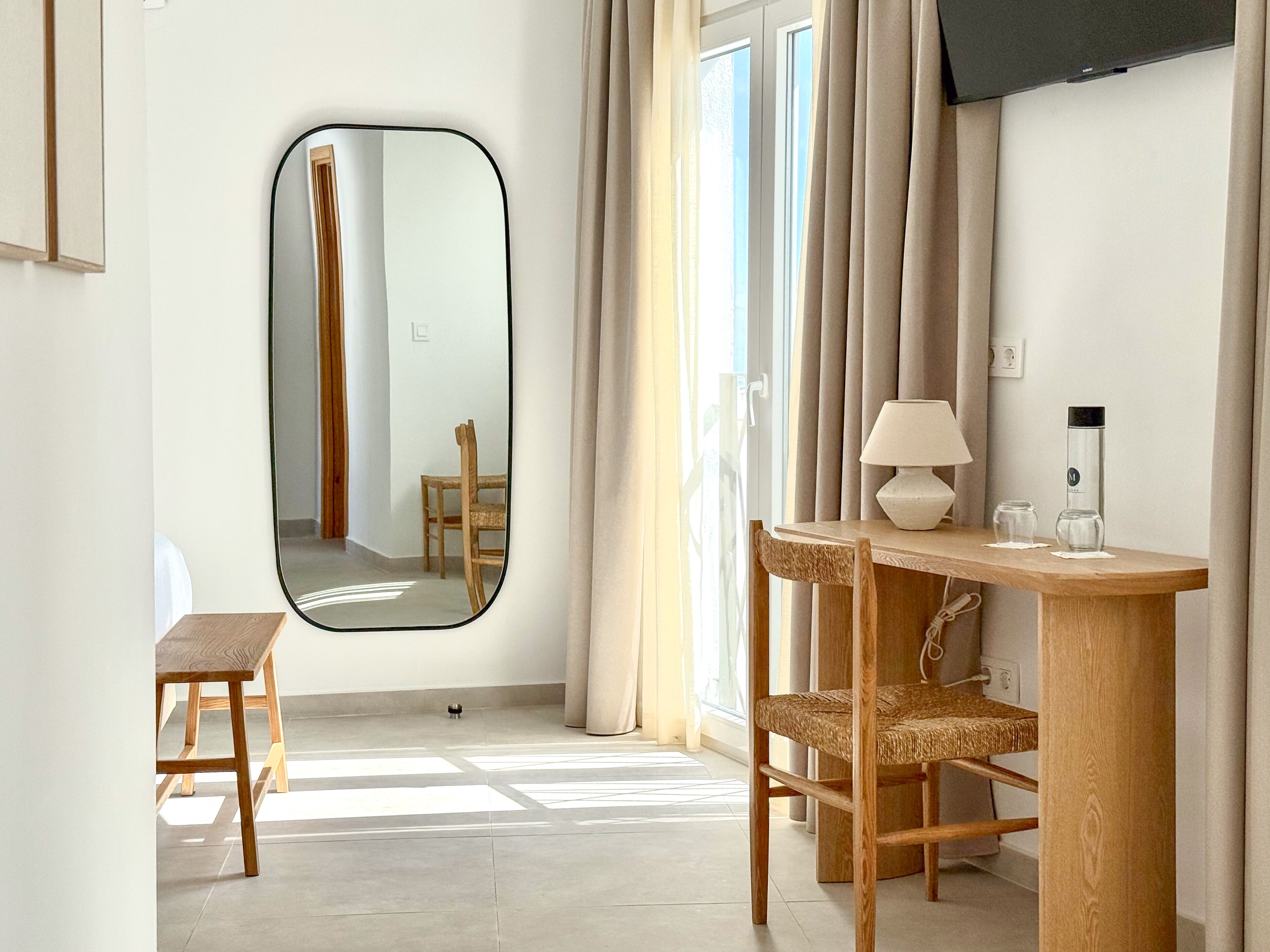 Una habitación moderna con un espejo grande, un escritorio de madera, dos sillas de mimbre, una lámpara y un televisor en la pared.