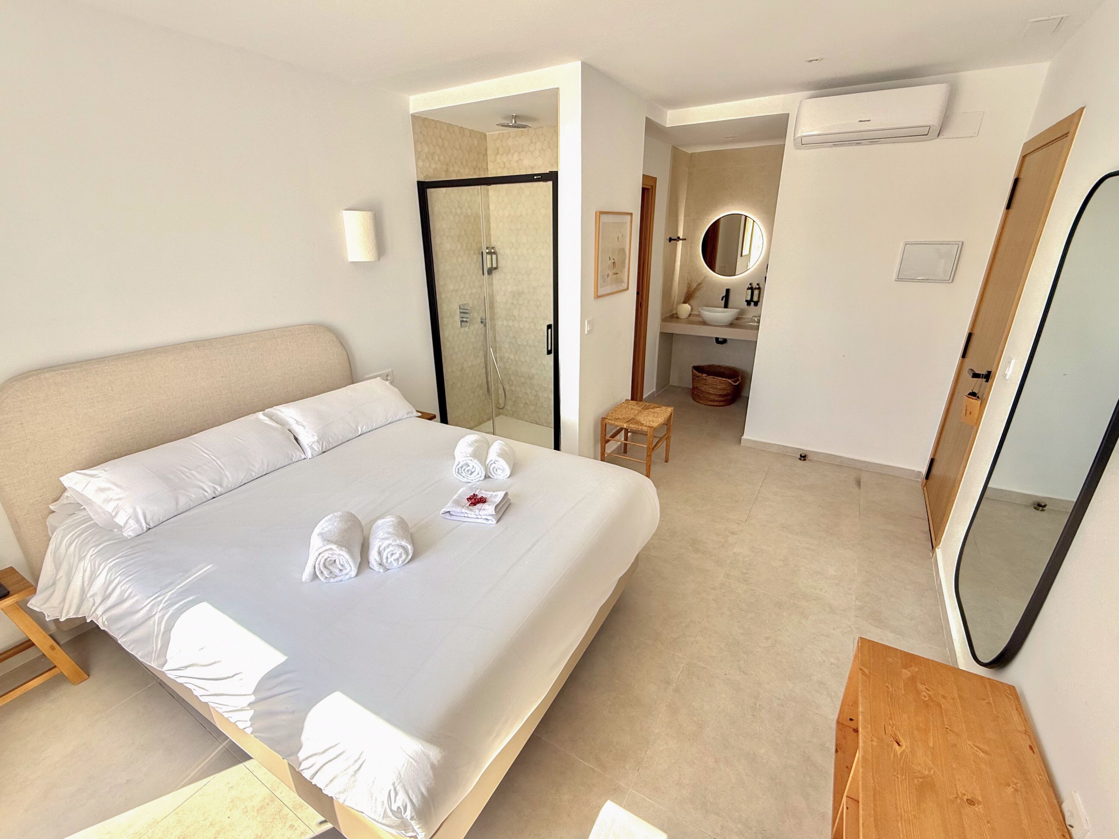 A hotel room with a bed, a wooden nightstand, a bathroom with a mirror, and a shower.