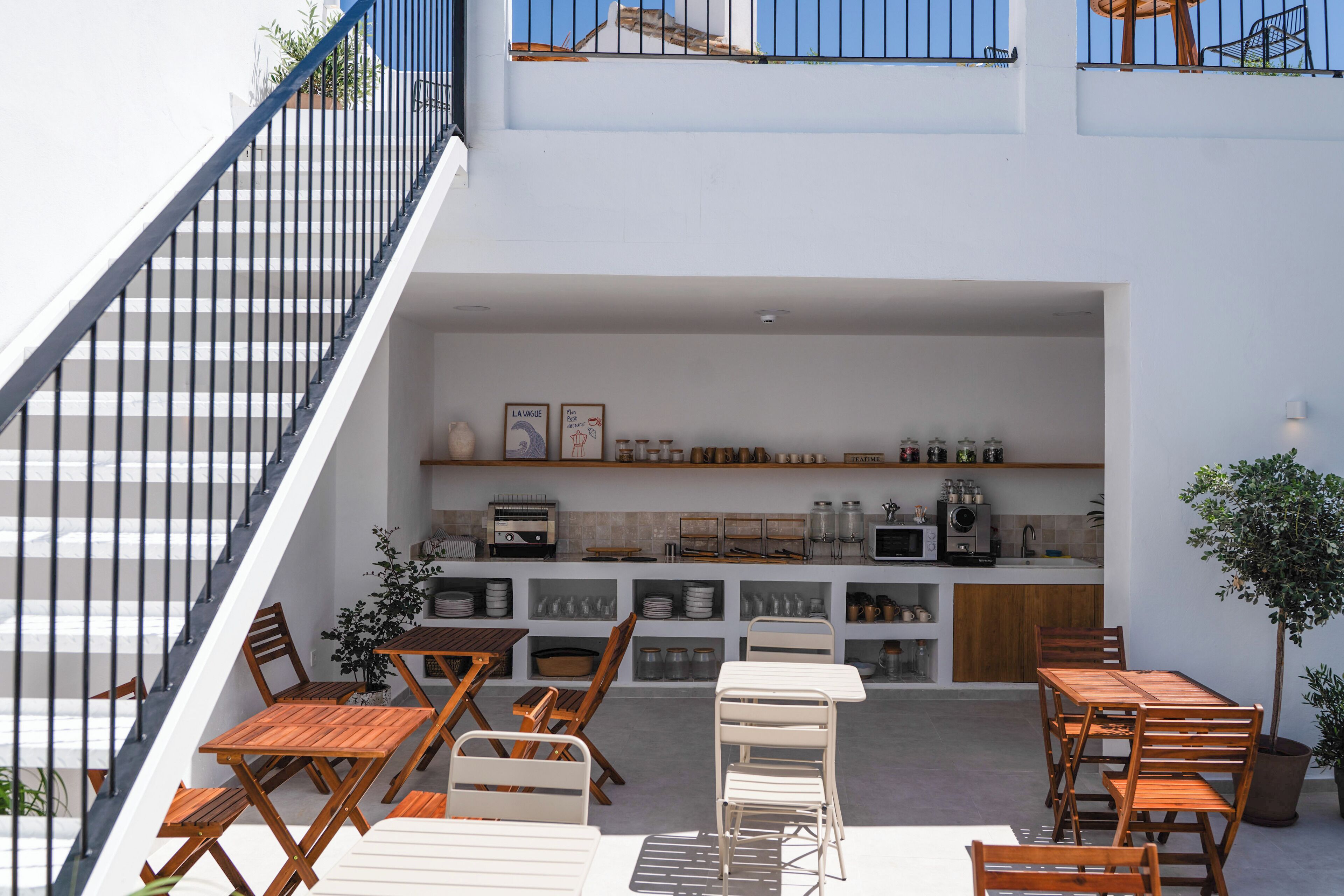A modern outdoor dining area with wooden tables and chairs, a kitchen area with shelves, and a staircase.
