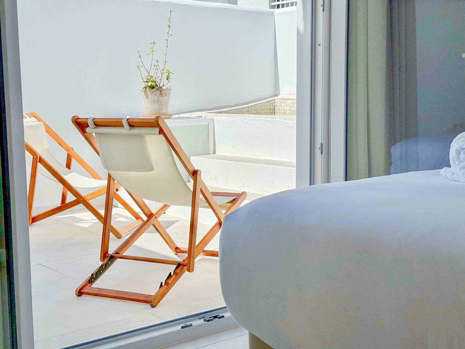 A bedroom with a bed, a wooden chair, and a potted plant on a balcony.