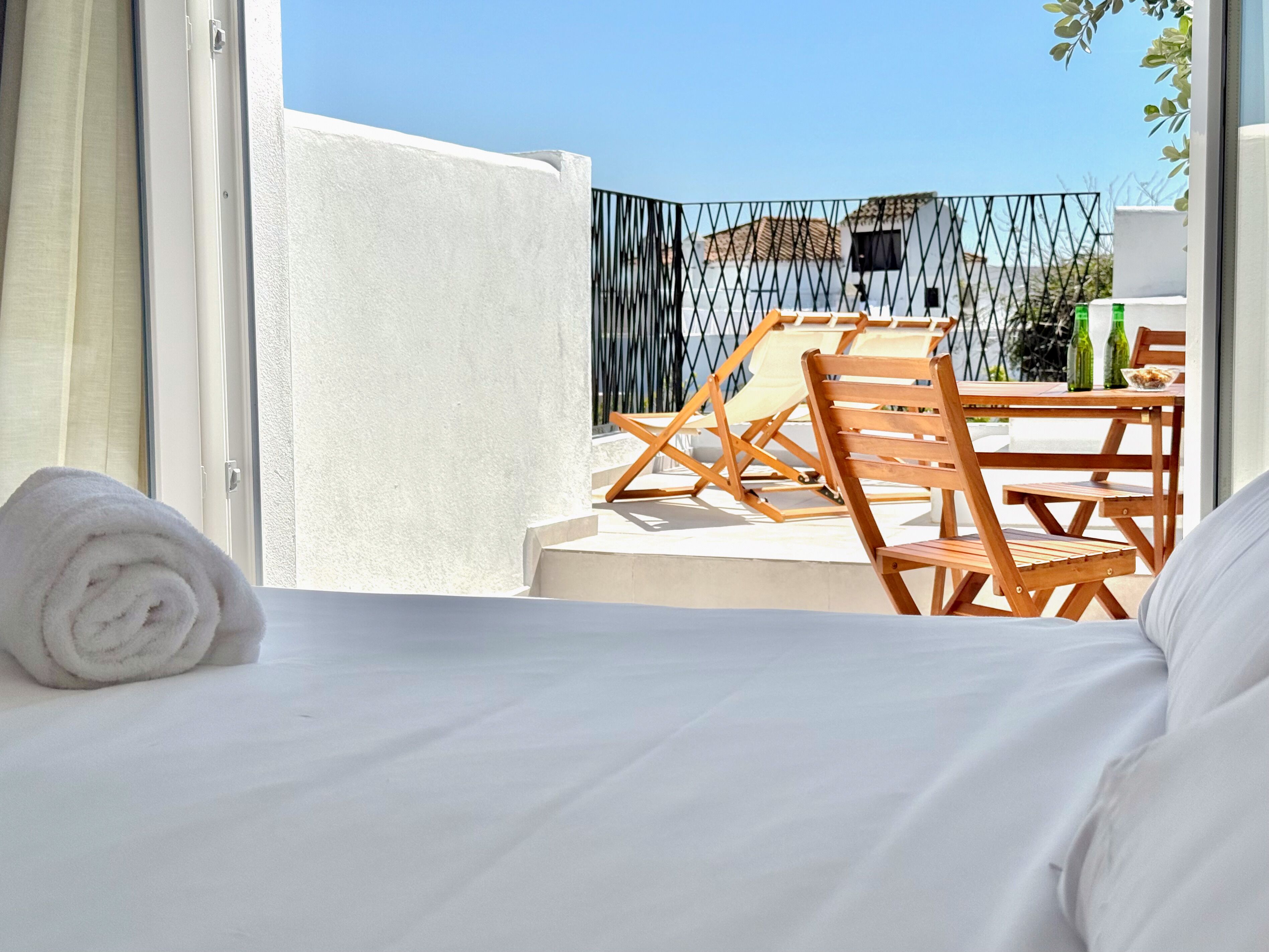 Habitación de hotel con cama, ventana, balcón con sillas y mesa de madera, y vistas a edificios.