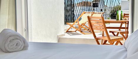 Habitación de hotel con cama, ventana, balcón con sillas y mesa de madera, y vistas a edificios.