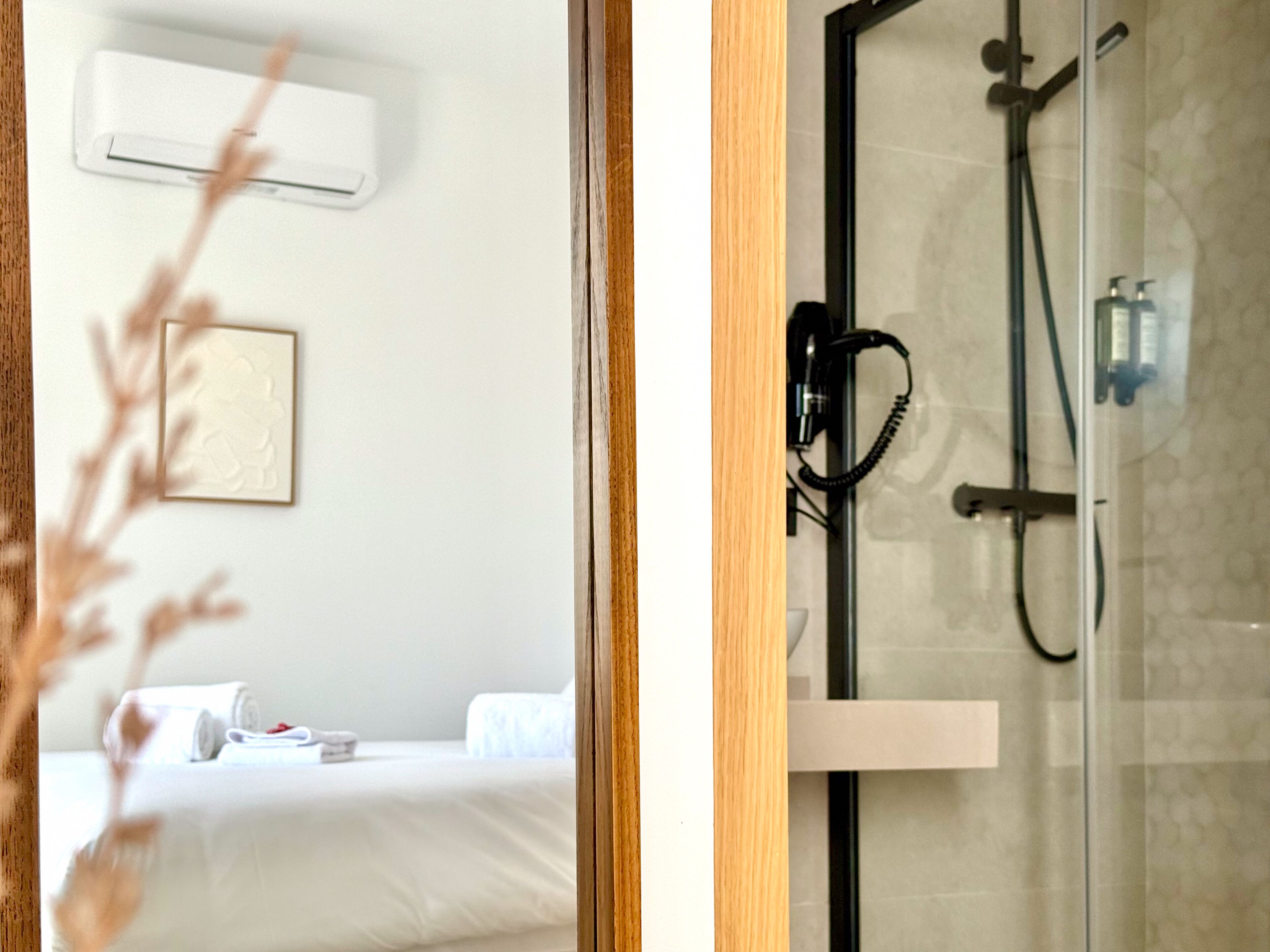 A hotel room with a bed, a bedside table, an air conditioning unit, and a shower area with a glass door.