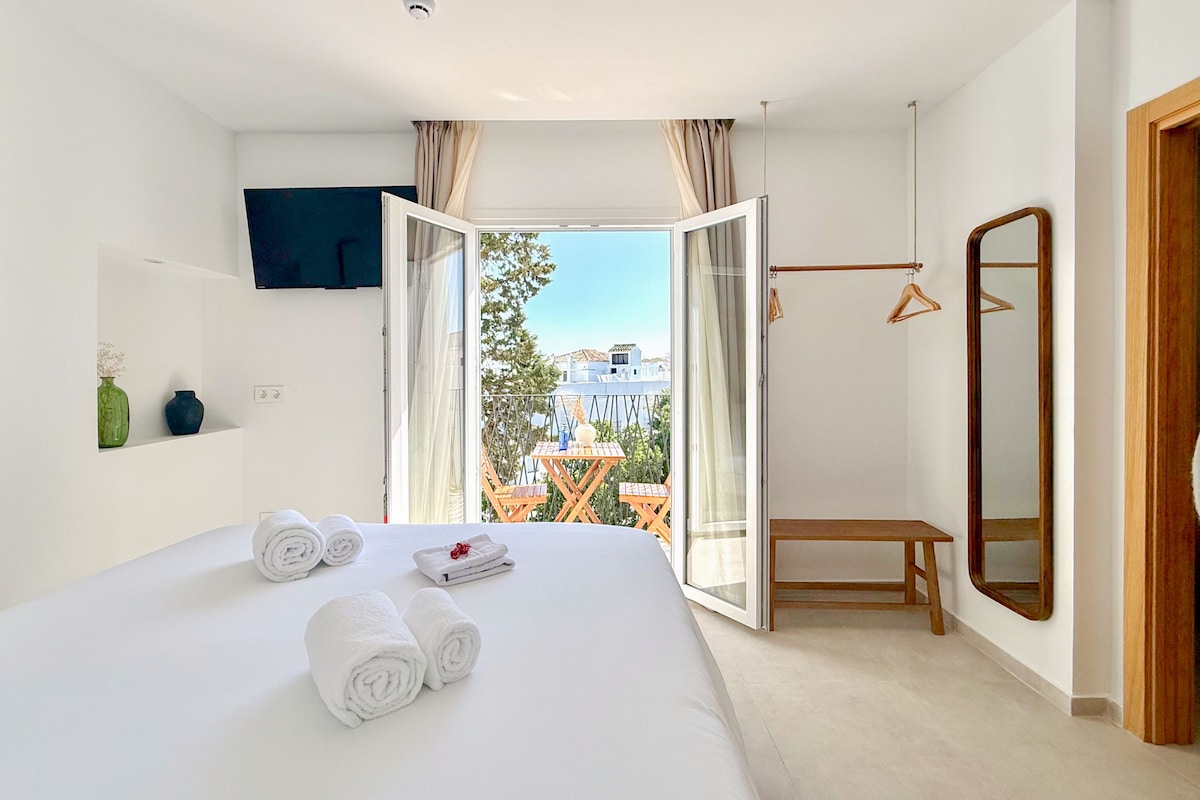 Una habitación de hotel con cama, televisión, balcón con mesa y sillas, y un banco de madera.