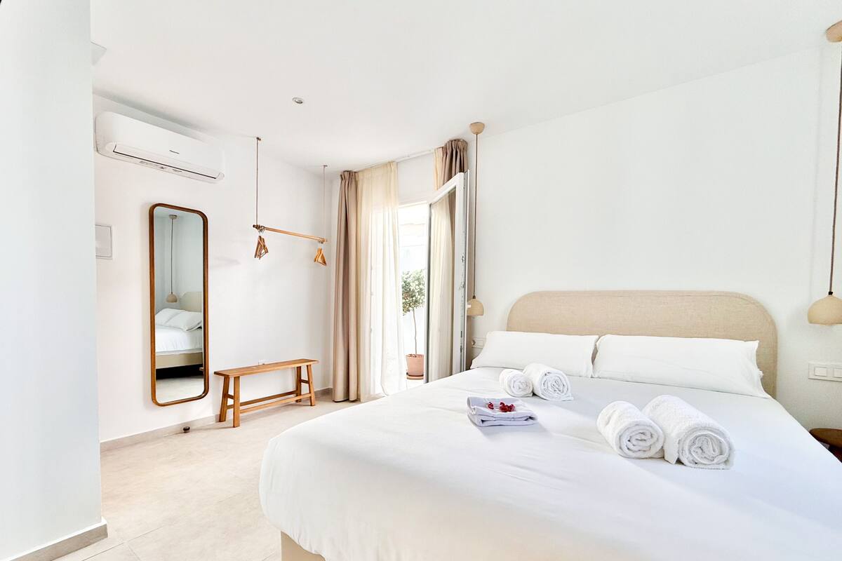 A hotel room with a bed, a bedside table, a mirror, a bench, and a window with curtains.