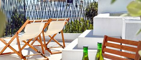 Terraço no telhado com vista para o mar, duas cadeiras de madeira, uma mesa e duas garrafas.