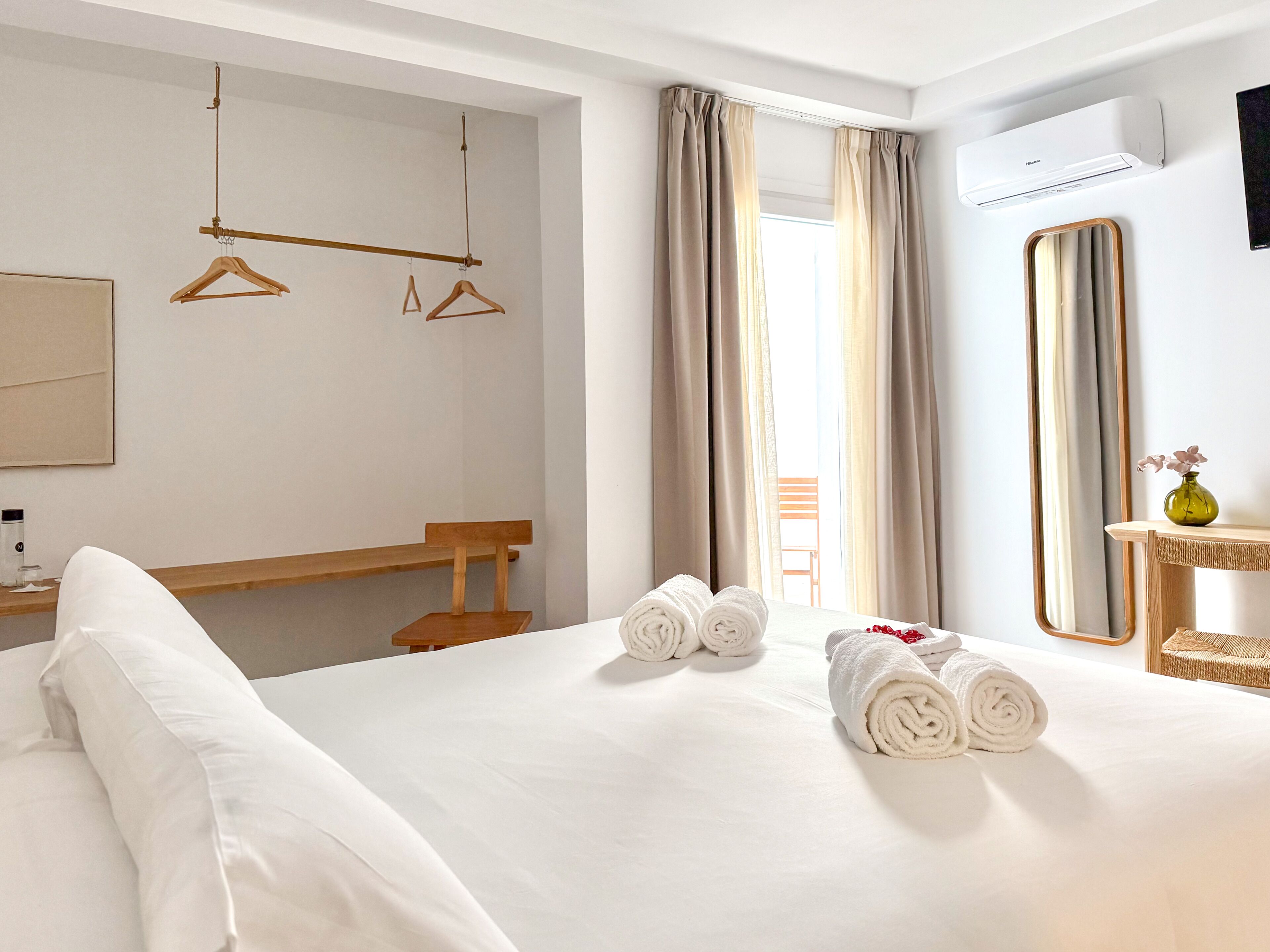 Une chambre d’hôtel avec un lit, un banc en bois, un porte-vêtements suspendu, un miroir et une fenêtre avec des rideaux.