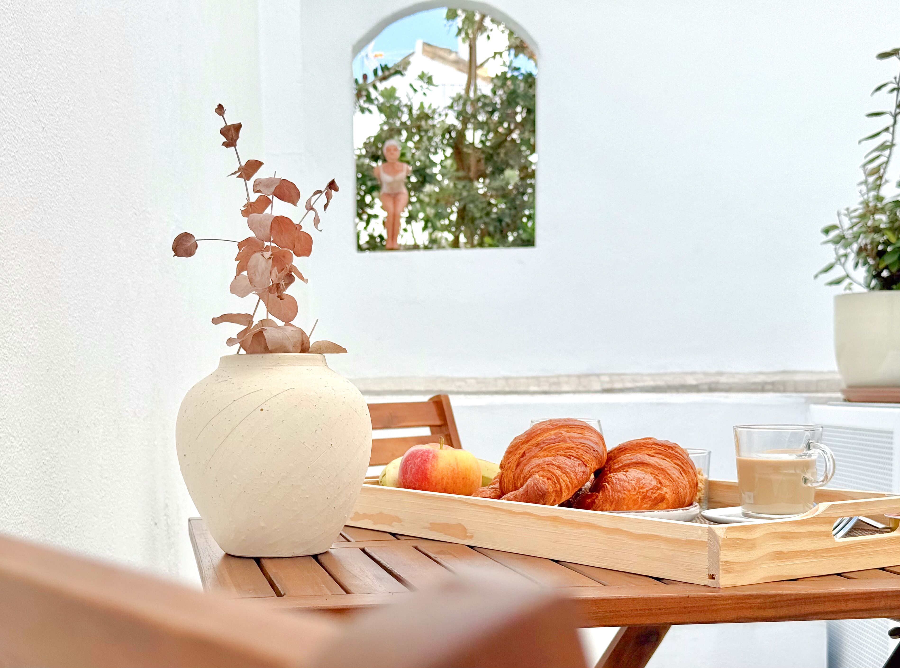 Una mesa con un jarrón de flores secas, una bandeja con croissants y fruta, y una taza de café.