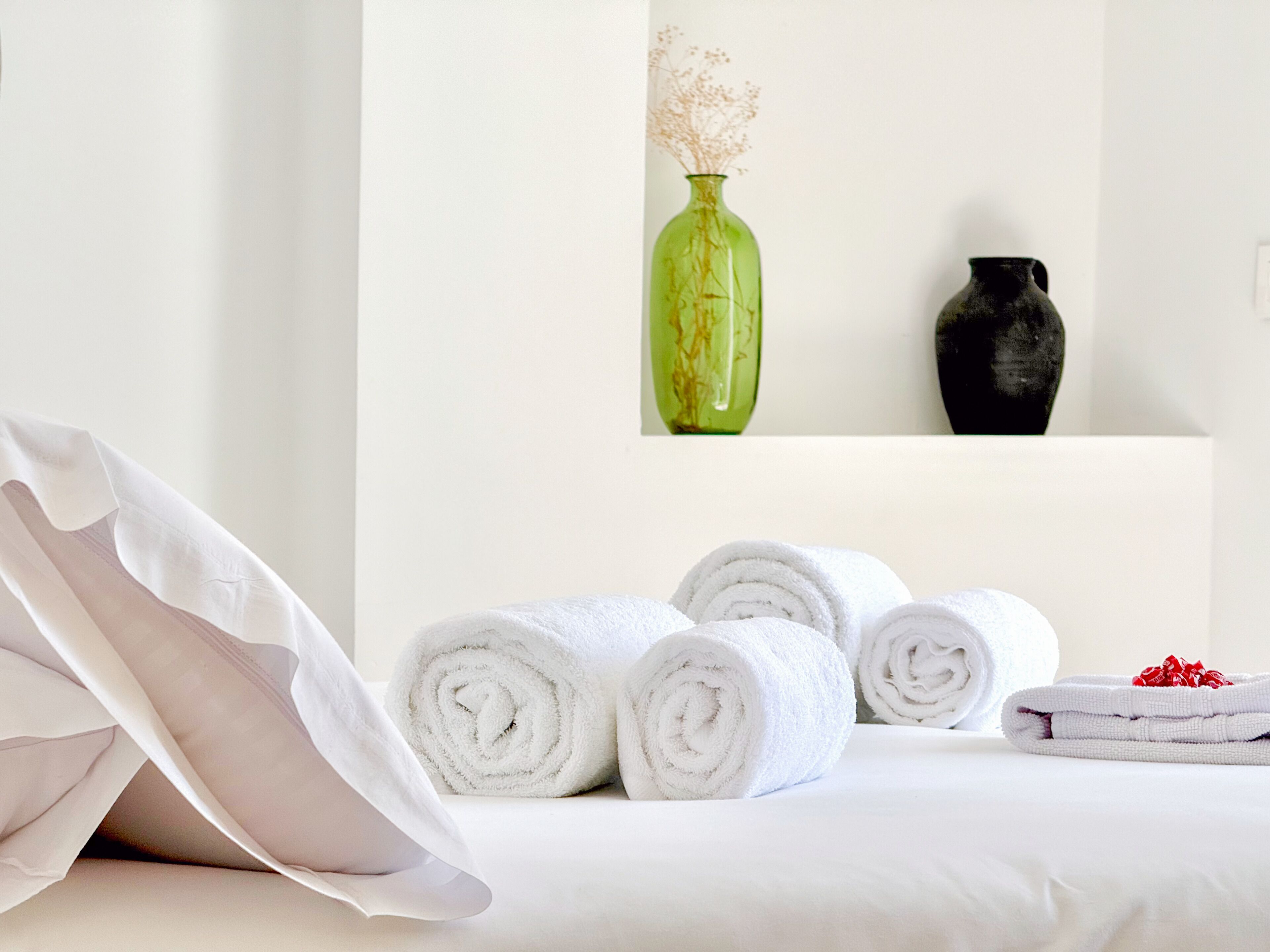 Una cama bien hecha con toallas blancas enrolladas, una almohada y dos jarrones sobre un estante blanco.