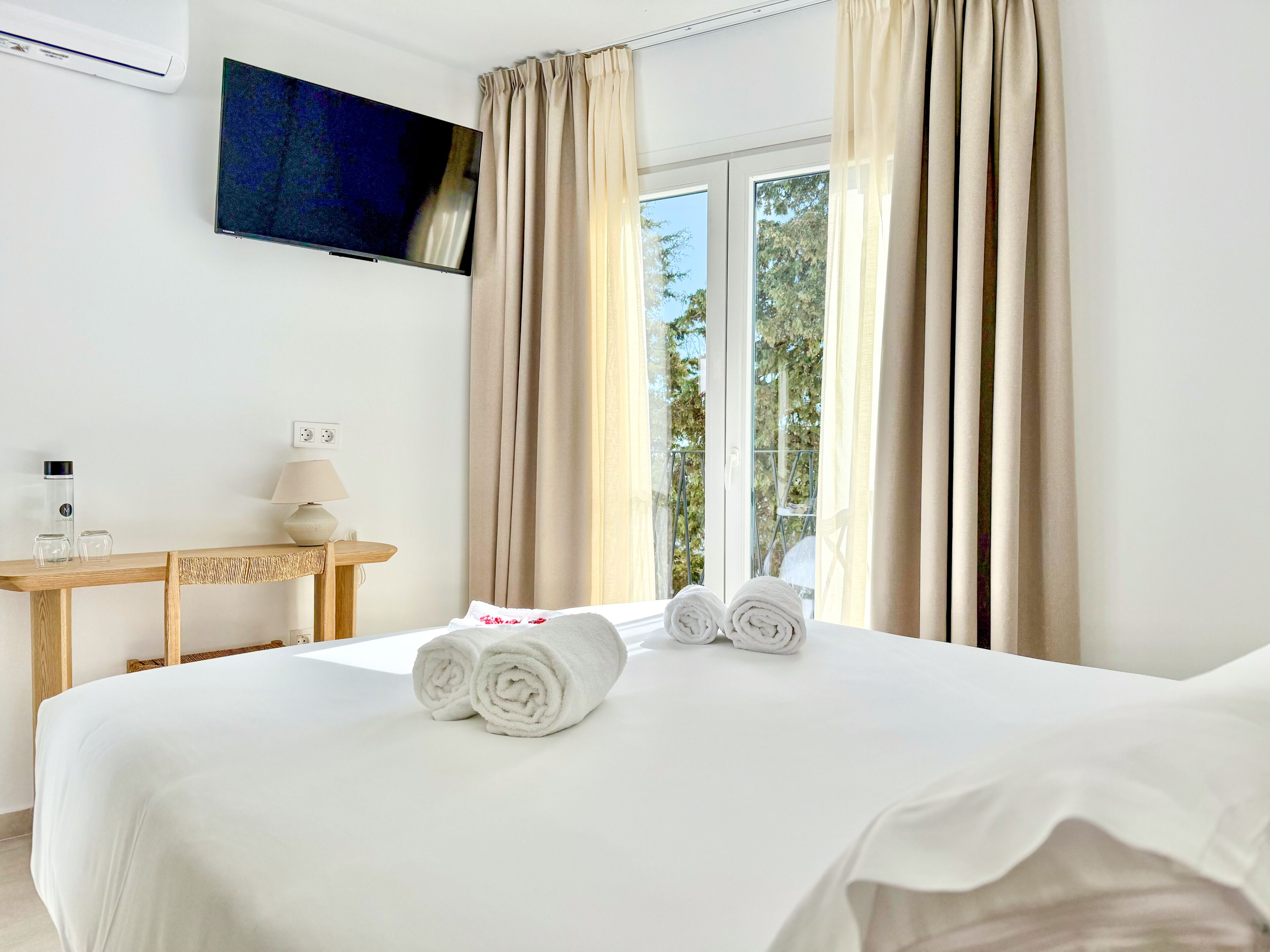 Habitación de hotel con cama, televisor montado en la pared, escritorio con lámpara y ventana con cortinas.