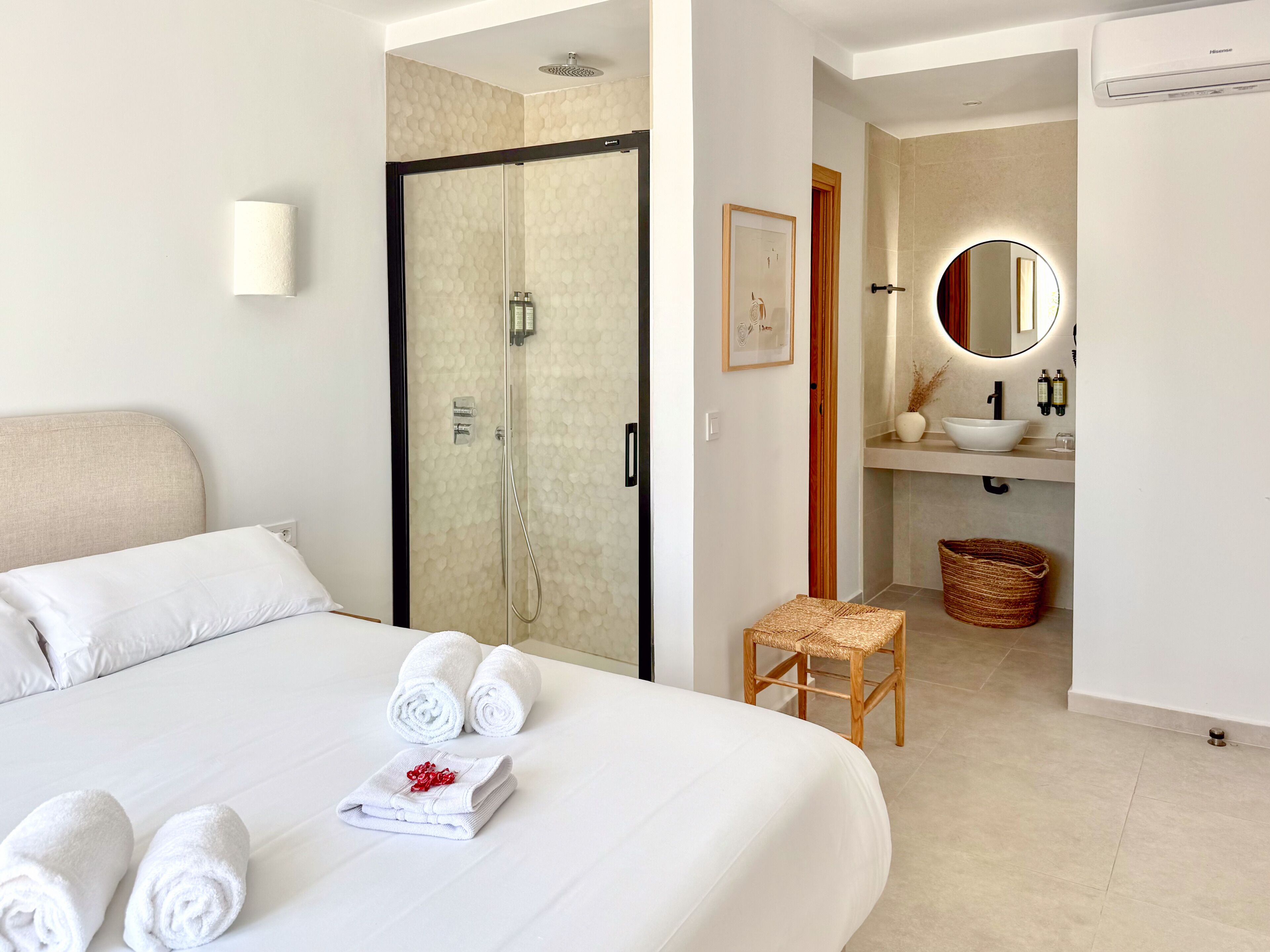 Habitación de hotel con cama, ducha, baño con lavamanos y espejo, y un taburete pequeño de mimbre.