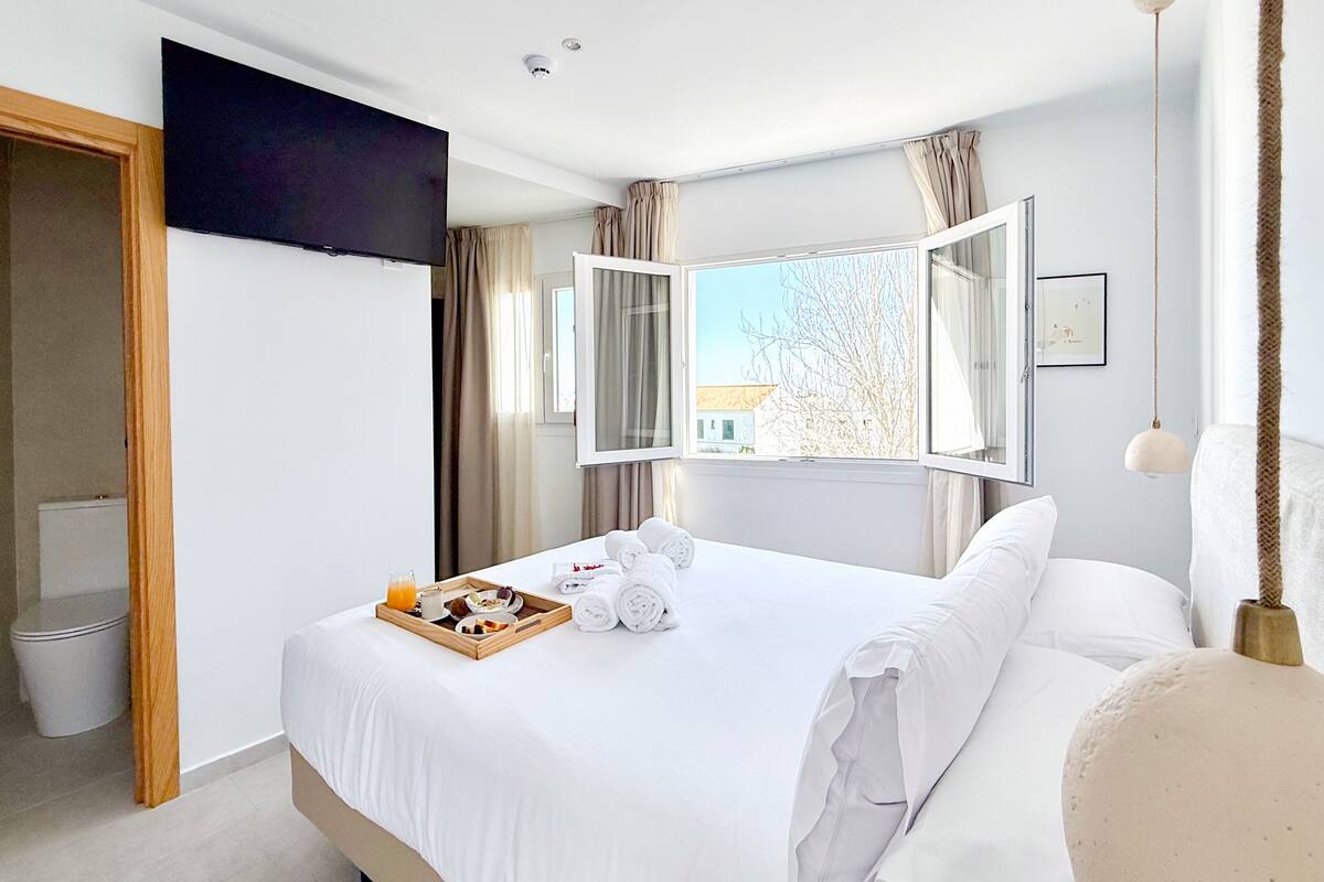 A hotel room with a bed, a tray with breakfast items, a TV, a window with curtains, and a view of the outside.
