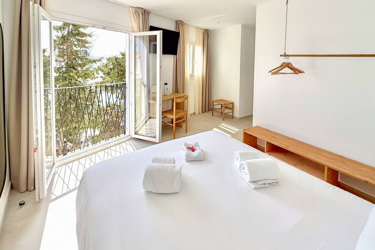 A hotel room with a large bed, a wooden bench, a small table, and a balcony with a view of trees.