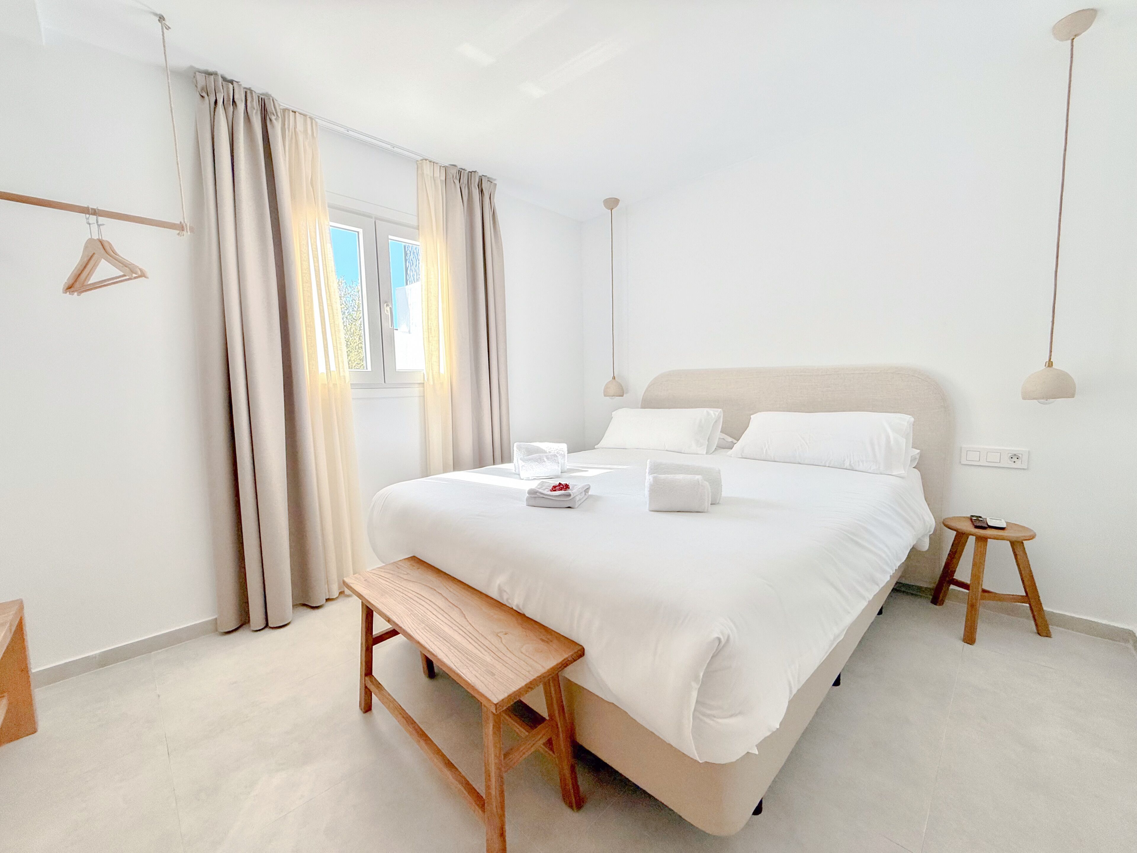 Quarto de hotel minimalista com uma cama grande, mesas de cabeceira, um banco e vista para o exterior.