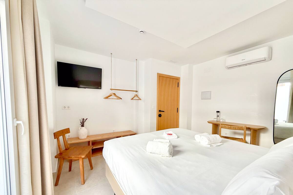 A hotel room with a bed, a desk, a chair, a TV, a door, and an air conditioning unit.