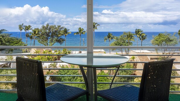 Een balkon met een glazen tafel en twee rieten stoelen, met uitzicht op een strand met palmbomen en de oceaan.