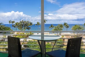A balcony with a glass table and two wicker chairs, overlooking a beach with palm trees and the ocean. - Kamaole Beach Royale 506 - Stunning Ocean Views, Spectacular Renovation, AC (Kihei)