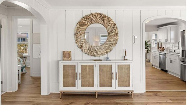 Un couloir aux murs recouverts de panneaux blancs, avec un miroir à motifs tressés au-dessus d’un meuble en osier, qui mène à un espace cuisine.