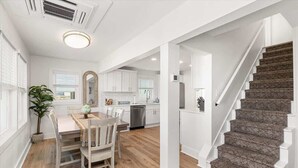 Une cuisine moderne avec des meubles blancs, un coin repas comprenant une table et des chaises, et un escalier recouvert d’une moquette à motifs.