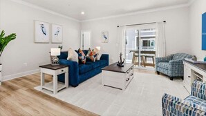 Un salon moderne avec un canapé bleu, une table basse blanche et des fauteuils à motifs. Des œuvres d’art encadrées ornent les murs, et une porte vitrée offre une vue sur l’extérieur.