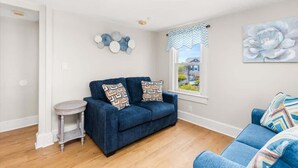 A living room with a blue sofa, a small round table, and a window with a view of buildings. - Dorchester Drift B: Shore Nest (Ocean City)