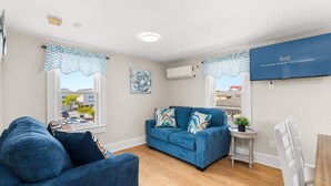 A living room with a blue sofa, a white armchair, a small side table with a plant, and a TV mounted on the wall. - Dorchester Drift B: Shore Nest (Ocean City)