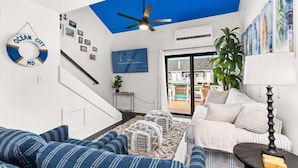 A living room with a blue ceiling, a sofa, ottomans, a lamp, and a wall with framed pictures and a life preserver. - Blue Horizon at Old Port (Ocean City)