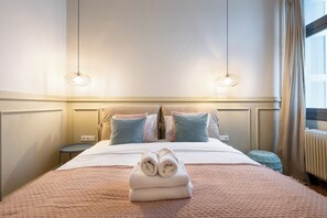 A neatly made bed with a folded towel in the center, a bedside table, and a window with curtains. - Meadows - Luxury 3-Bedroom Apt in Athens (Athens)