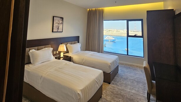 A hotel room with two beds, a desk, a TV, and a large window with a view of the sea. - Hams Apartments (Duba)