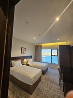 Habitación de hotel con dos camas, un escritorio, un televisor y un ventanal con vista al mar.