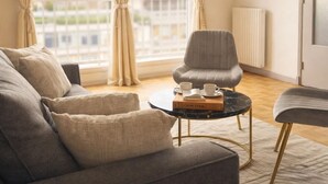 Un salon moderne avec un canapé gris, une table basse ronde sur laquelle se trouvent un livre et deux tasses, et une vue sur le paysage urbain à travers de grandes fenêtres.