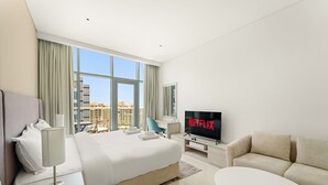A modern hotel room with a large bed, a TV displaying 'NETFLIX', a sofa, and a view of the cityscape through large windows. - Seven Palm - 1421 Studio (Dubai)