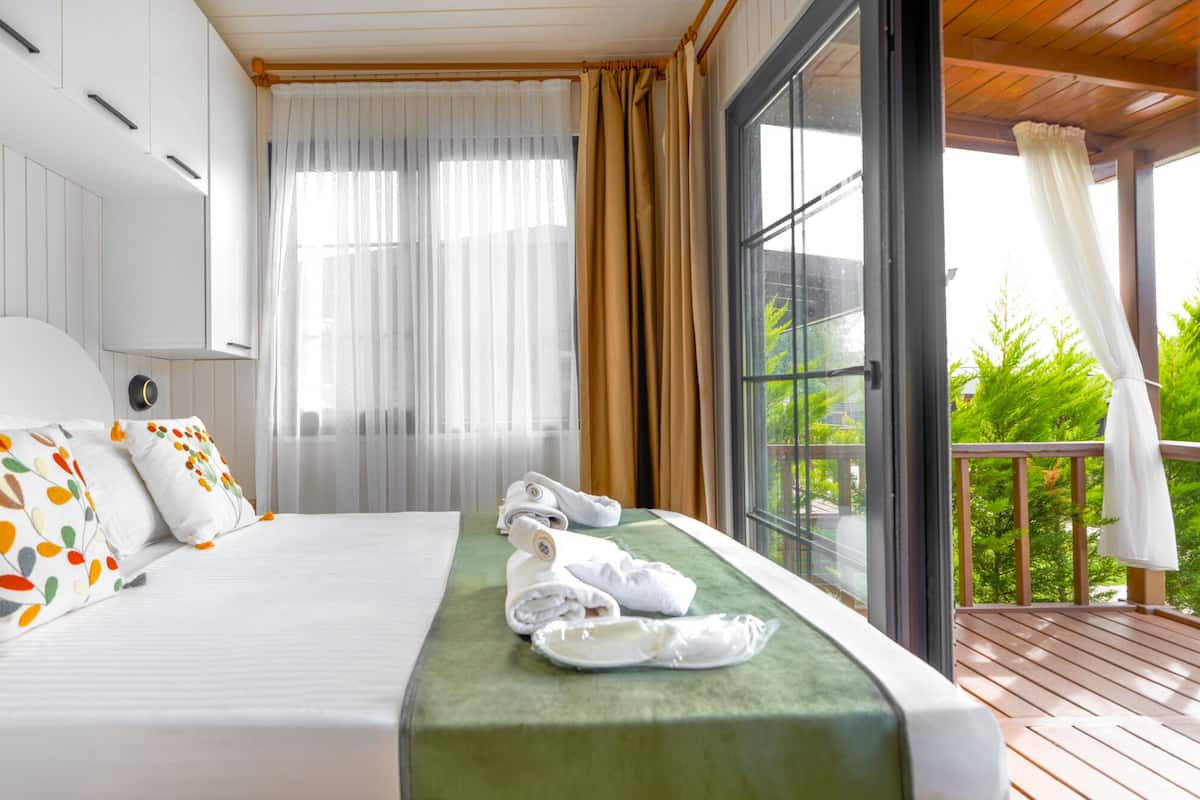 A hotel room with a bed, a green bedspread, white pillows, a window with curtains, and a balcony with a view of greenery.