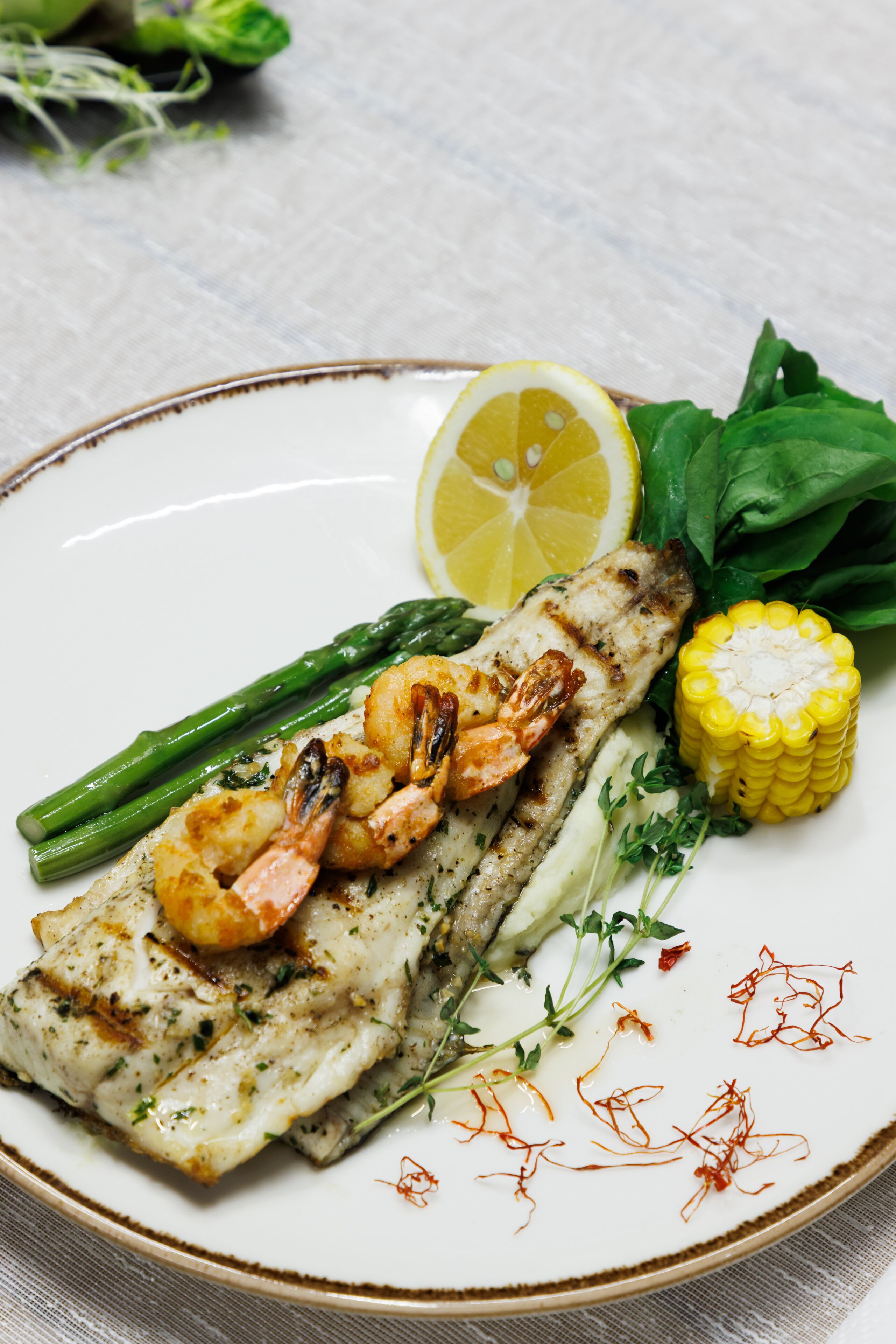 Un plato con pescado a la parrilla, camarones, espárragos, maíz y limón.