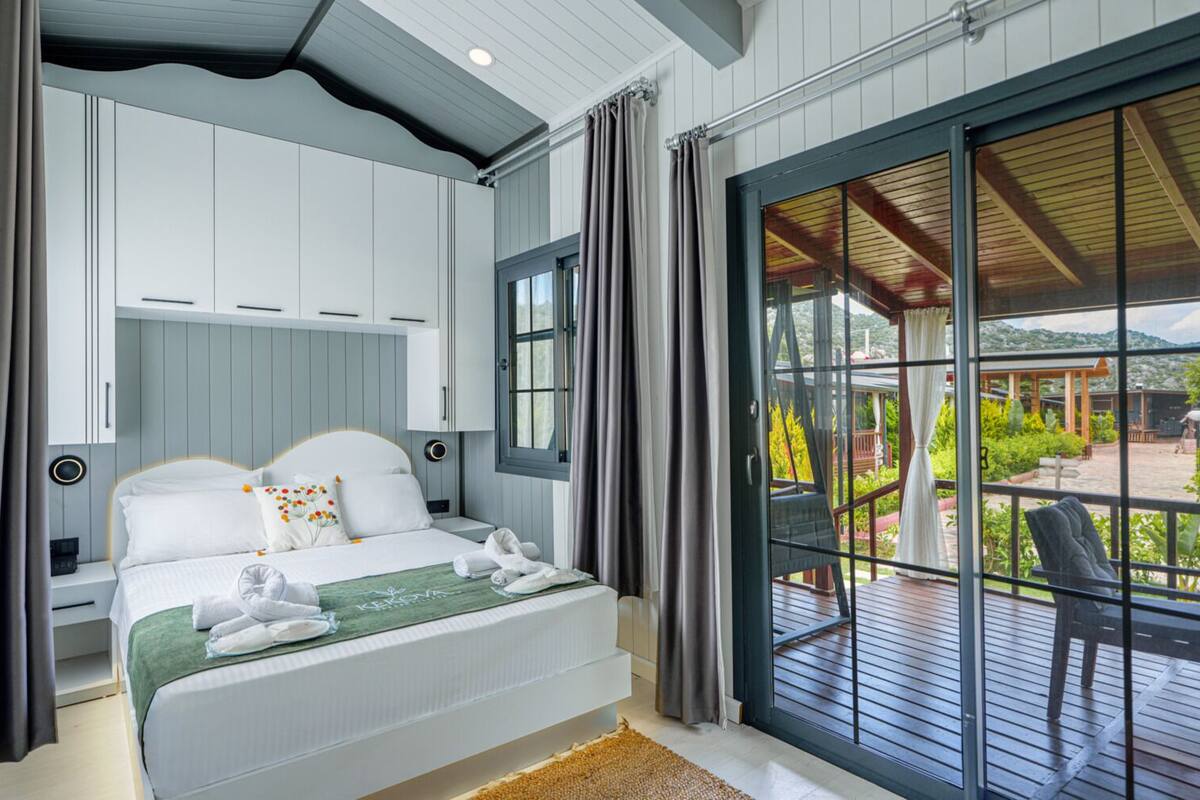 A bedroom with a bed, a nightstand, a window with curtains, and a view of an outdoor deck.