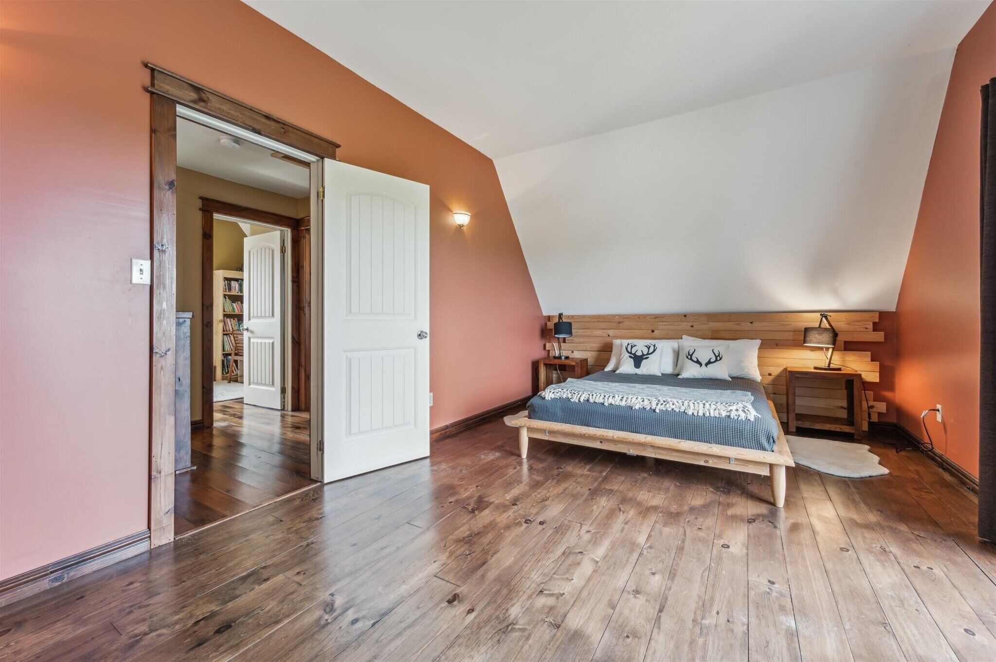 A bedroom with a sloped ceiling, wooden flooring, a bed with a headboard, a nightstand, and a wall-mounted lamp.