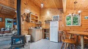 Interior acogedor de una cabaña, con paredes de madera, una estufa, un refrigerador, una mesa de comedor con sillas y una ventana con vistas a árboles.